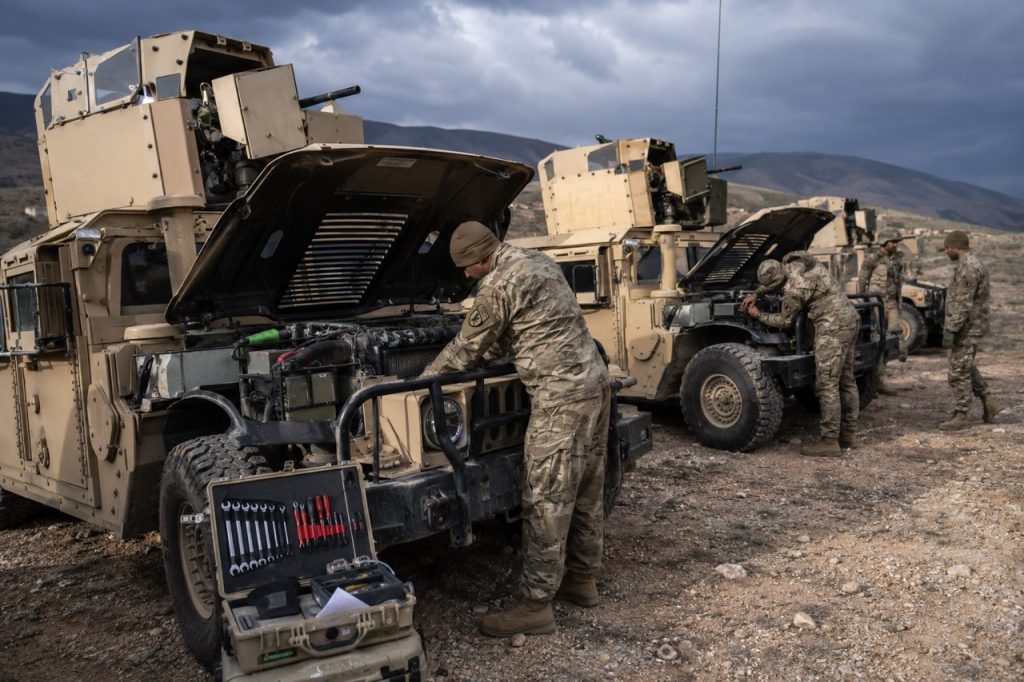 Mecânica militar realizando manutenção de campo em veículos blindados HMMWV em um ambiente deserto robusto com compartimentos de motores abertos e equipamentos táticos.