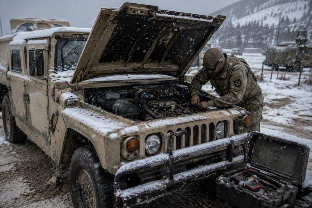 A military maintenance technician repairs damaged electrical wiring inside a Humvee during a field operation in harsh weather conditions.